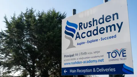 Northamptonshire Police, Fire and Crime Commissioner A school sign, written in blue and white, by a large tree. There is writing on the sign. 