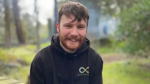 David Smith has short brown hair and a short beard. He is smiling and wearing a black hoodie that has Neurotribe written on the right with an infinity symbol. He is sitting in the woodland with grass and trees around him.