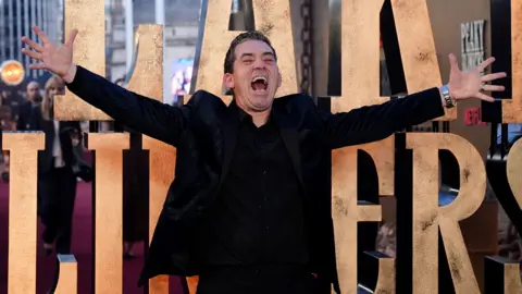 PA Media A man with outstretched arms and a big smile stood in front of a gold Peaky Blinder sign. He has black hair and black jacket and shirt.
