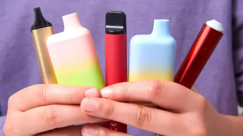 Two hands that look young holding five vapes which are brightly coloured.
