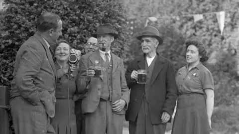 Black and white photo shows BBC's Ralph Wightman (left) recording a toast to victory at the public house at Piddletrenthide, Dorset. His is holding a large microphone with a large BBC logo on the top. He is with three men and two women. Two of the men are wearing suits and trilby hats and holding up pint mugs. The recording formed part of Victory Report in BBC programmes on VE night.