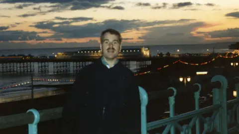 John Reidford A man stands with a beach and a pier behind him as the sun sets. 