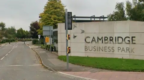 The entrance to the Cambridge Business Park. On the right is a stone wall with Cambridge Business Park written on it in silver letters. There is a traffic light in front of it. The road stretches down and has small trees. 
