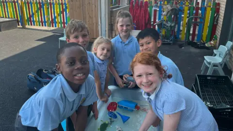 Three boys and three girls cluster around a water tray outdoors with plastic toys floating in it. They are in the shade and smile at the camera. They wear pastel blue uniform. Behind them is a colourful fence.