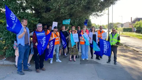 People on a picket line. There is a mixture of men and women and they have flag and posters with their unions' names on them. It is a sunny day and they are standing on a concrete drive with trees in the background