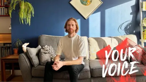 BBC Male sitting on sofa wearing T shirt and dark trousers in own home with blue walls and grey sofa