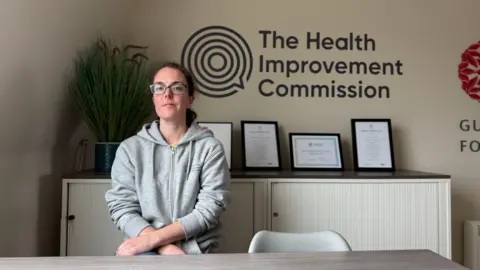 A women in her mid 30s with glasses and her brown hair tied back in a grey zip up sweater sat on a stool in front of a cabinet with a green plant behind her right shoulder and certificates on her left. The wall has the logo of the Health Improvement Commission on the wall above the certificates.