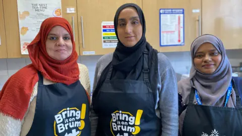 Three women are wearing black aprons that say 'Bring it on Brum'. They are wearing different coloured hijabs and smiling while standing in a kitchen.