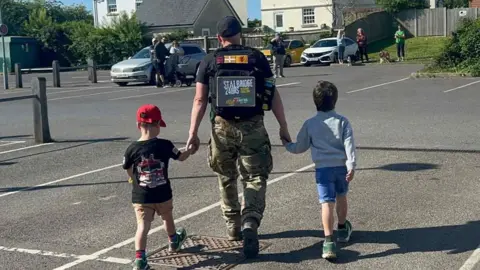 Francesca Harris Martyn Harris has is back to the camera and his holding his two young sons hands. Martyn is wearing a black t-shirt advertising his Stalbridge 24 hours walking challenge and army trousers. His son on the left has a red cap on, black t-shirt and brown shorts while the one on the right has blue shorts and a white jumper.