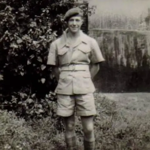 Family photo A black and white photo of a young Peter Byers standing in a West African village. He is wearing a beret, khaki shorts, belt and short-sleeved army uniform, long socks and boots. He is smiling at the camera.