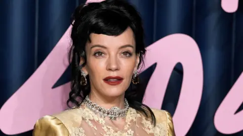 Lily Allen, with dark hair pinned up, wearing a gold dress, in front of large pink writing.