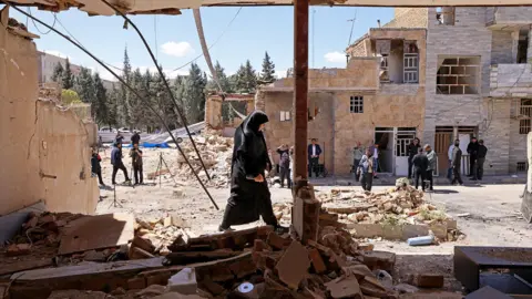 AFP via Getty Images A woman walks past the residential buildings that were damaged by recent strikes at Vahdat town in Karaj, southwest of Tehran on 3 April, with bystanders and other buildings in the background against a sunny blue sky.