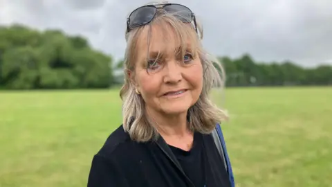 JOANNA TAYLOR/BBC Diane Harvey is looking at the camera. She is wearing a black top and has sunglasses on top of her head. Behind her is a green field. 