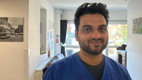 Mukesh smiling at the camera. He has short black hair and short stubble and wears blue care home scrubs with a grey layer underneath. The white walls behind him have artwork on including a black and white street scene, and there are large windows behind him.