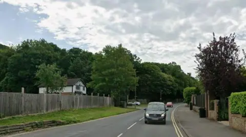 A leafy road with cars driving on it and a white house in the background.