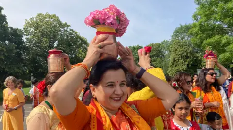 A smiling woman wearing an orange sari holds a gold bowl full of pink flowers above her head