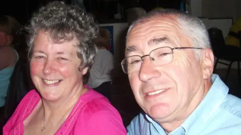 Family A couple pictured in a room with others in the background. They are both smiling. The woman is wearing a pink scoop-necked t-shirt and has light curly short hair. The man is in a pale blue shirt and wire-framed glasses. He has white hair swept back from his forehead.