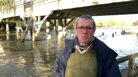 Neil Debono is wearing a blue fleece over a green hoodie. He has short brown hair and is wearing glasses. Behind him is a wooden bridge over the river Thames.