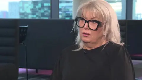 Baroness Helen Newlove is looking away from the camera while being interviewed. She has long blonde hair and is wearing large black-framed glasses and a black top with a microphone fixed to the collar.