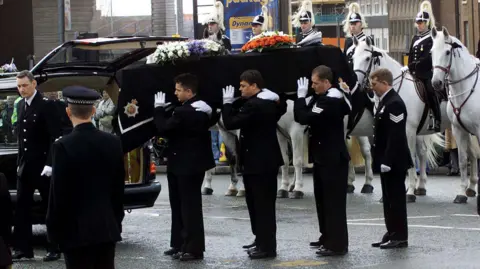 Getty Images The coffin of Detective Constable Stephen Oake is held by six pallbearers all in Greater Manchester Police uniform with white gloves. In the background mounted police officers have formed a guard of honour for the funeral cortege.