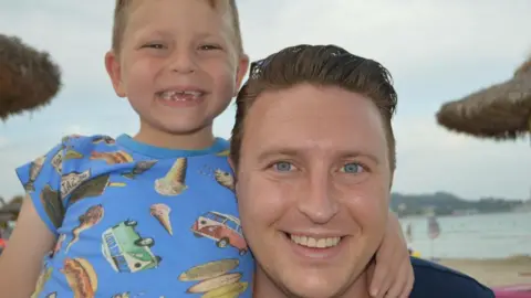 Image of a young boy with a blue t-shirt with images of camper vans and ice creams with his arm around a man with blue eyes and short brown hair. The boy has gaps in his teeth but he and the man are smiling at the camera. In the background can be seen sand, sea and beach umbrellas.