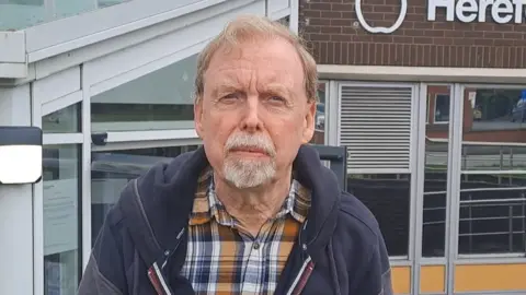 Councillor Louis Stark, a man with light brow hair who is wearing a yellow checked shirt with a navy blue jacket. He is standing outside a council building with glass entrance and railings.