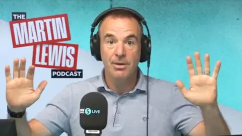 Martin lewis has short day hair, is wearing headphones and a blue polo-shirt and had his hands up to the side of him.