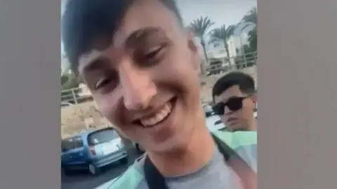 Handout A man with short dark hair smiles at the camera. He is taking a selfie and is wearing a light blue t-shirt
