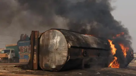 Burning wreckage of a fuel tanker, which lies on its side by the roadside