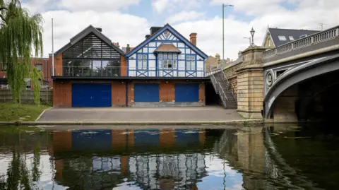David Johnson Photographic A boathouse on the bank of a river is mainly blue and white. There is a bridge next to it.