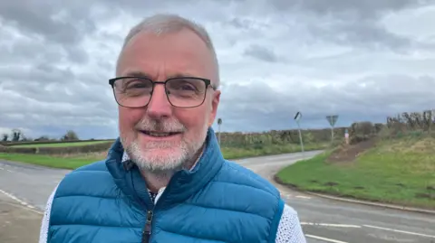 John Breckon is standing by the B1257 in North Yorkshire. He has short grey hair and a short grey beard and is wearing a blue gillet over a pale shirt