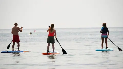 Two women and one man are standing up on paddleboards with oars in their hands. The man is wearing only shorts, the woman in the middle is wearing a vest top and shorts and the woman to the right is wearing a rash vest and shorts.