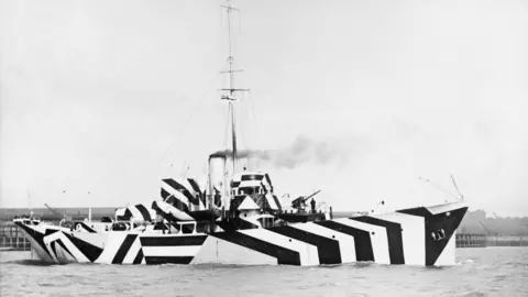 A black and white image of a large wartime ship in water with smoke coming out of a funnel. It is painted with a black and white geometric stripe pattern