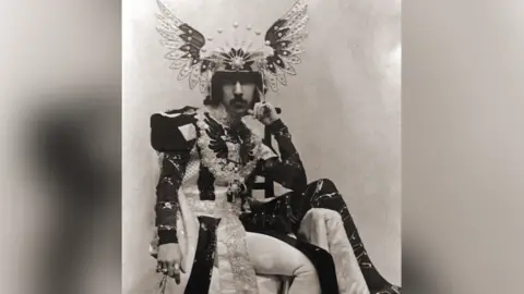 National Trust The 5th Marquess of Anglesey in a sepia photograph wearing a floor length overcoat consisting of differently coloured pieces of heavy material. The top of the coat is military in style with large shoulder pieces. He wears a headdress of splayed wings with a tiara-effect frontspiece.