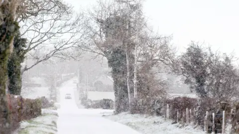 Pacemaker A white car is travelling on a snow covered road in the countryside. There are bar trees and bushes on both sides of the road. 