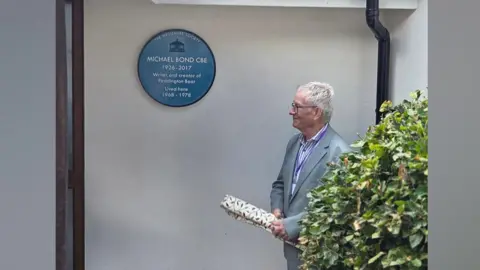 Dawn Brown Chris Harrison, chair of the Haslemere Society, unveiling the blue plaque at the former home of Paddington Bear author, Michael Bond. Chris is in a grey suit and is holding the curtain used to cover the plaque. The blue plaque is visible in the image.