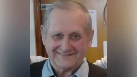 William Brown looking directly at the camera. He has grey hair. He is smiling. He is wearing a blue shirt, the collar of which can be seen, under a black jumper.