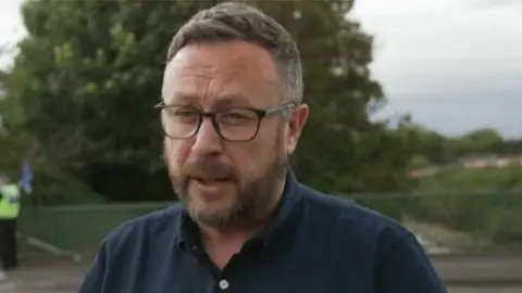 A man with glasses and a dark blue shirt, green railings and trees in the background, and a person wearing a safety vest