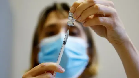 Reuters A needle going into a vaccine vial, a woman is holding both of them and is in the background. She is slightly blurred out but has blonde hair and a blue face mask on. 