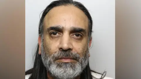 A police mugshot of a man with long dark hair, a grey beard and brown eyes.