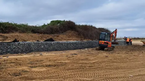 Tendring District Council A digger and workers on the beach at Walton-on-the-Naze. There is a long sea wall built of stones on the sand