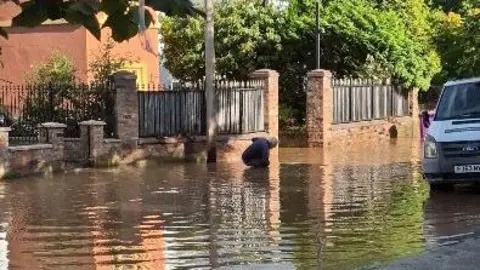 A street is flooded, and a man in crouching down in the water. There is a truck parked up on the side of the street.