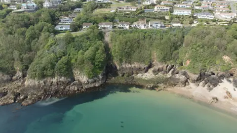 Soldier's Bay and the cliff face above it. At the top is Fort George a collection of large houses with brown roofs and white facades. Beneath the houses is a layer of greenery, with trees covering the middle half of the cliffs, above brown strips of granite getting darker as it goes closer to an azure blue sea. In the bottom left corner a brown patch of water is emerging from a small pipe into the sea. 