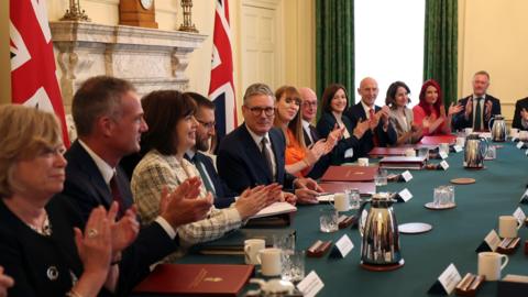 Sir Keir Starmer sits at a green table which has silver teapots and white mugs on it. The members of his cabinet are sat around the table and are clapping. Two Union Jack flags are in the background next to a white marble fireplace.