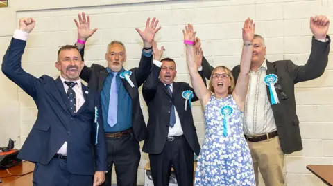 Five people wearing Reform UK rosettes raise their arms in celebration