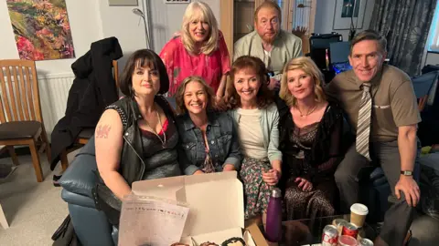 WHOCULT The Gavin and Stacey cast smile looking at the camera with a selection of custom donuts based on their characters faces sits in front of them.