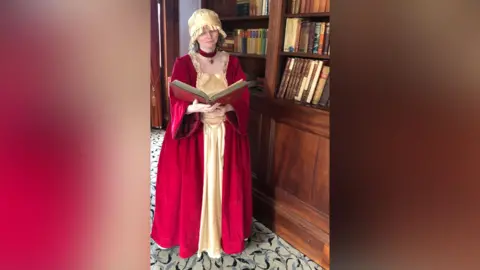 Mai Black A woman dressed in 18th Century ladies fashion. The dress is red with gold fabric in the middle. The woman wears a gold hat. She holds a large red book in her hands as she stands in front of a large wooden book shop.