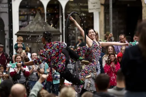 Ewan Bootman/Anadolu via Getty Images Male and female street performers in floral bright costumes pose on a metal ring prop with bubbles around them. People have gathered around them and are smiling and clapping. 