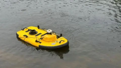 BBC/Charlotte Benton A bright yellow paddle board on a river. The water is dark and murky. The board has technical equipment on top which is used to monitor river health. 