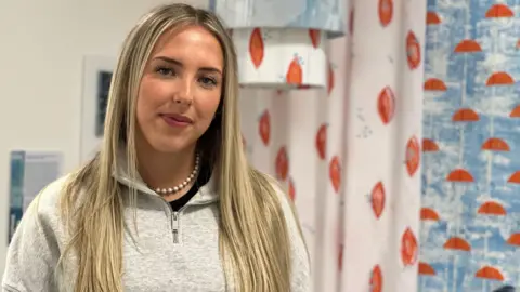 Sophie Boyde has long blonde hair and is wearing a grey top standing next to blue and orange designs on fabric.